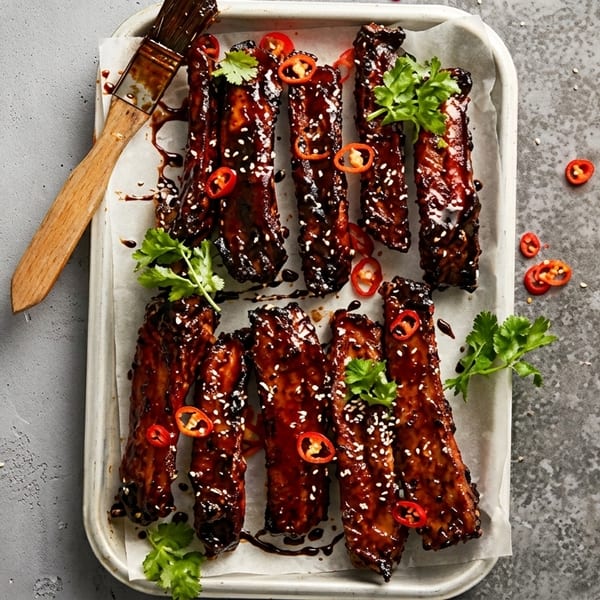 Glazed BBQ ribs with sesame seeds, chili, and cilantro on a white tray with a basting brush.