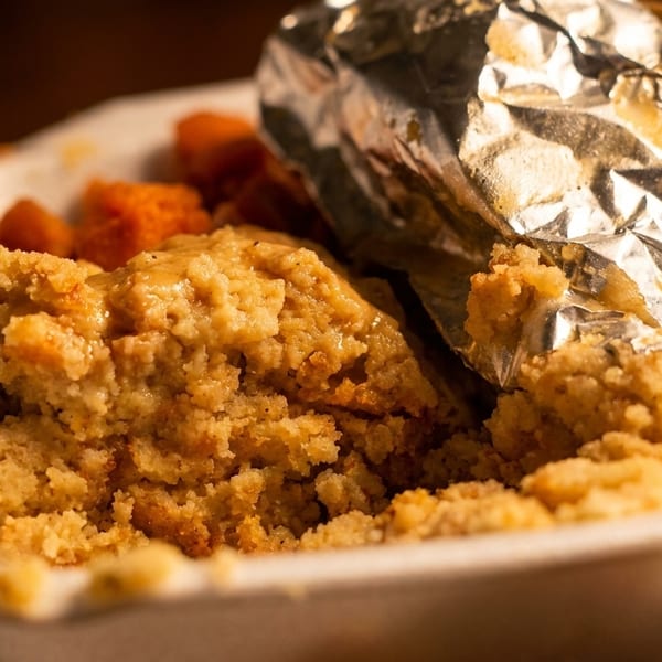 Takeout container with cornbread dressing, sweet potatoes, and a foil-wrapped baked potato.