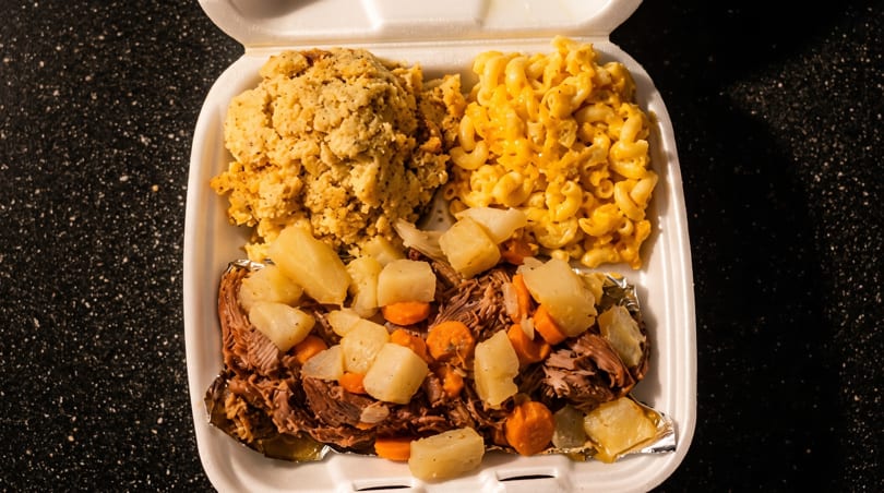 A takeout container with pot roast, potatoes, carrots, cornbread dressing, and macaroni and cheese.