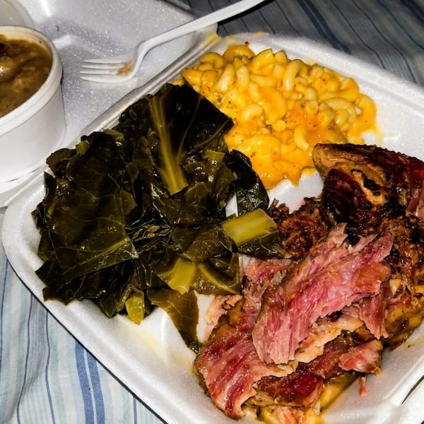 A takeout container with barbecue ribs, collard greens, macaroni and cheese, and a side of gravy.