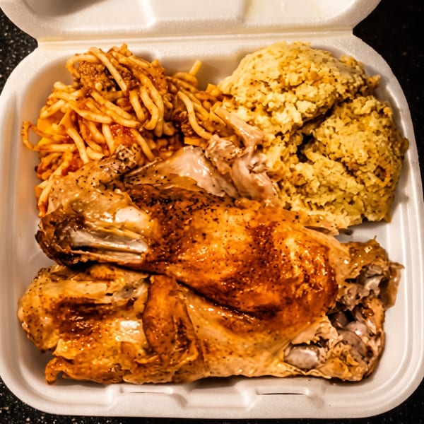 Roasted chicken, spaghetti, and rice in a white takeout container on a dark speckled surface.