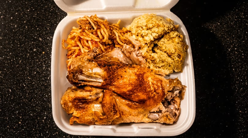 Roasted chicken, spaghetti, and rice in a white takeout container on a dark speckled surface.