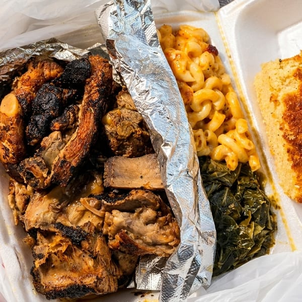 A takeout container with BBQ ribs, macaroni and cheese, collard greens, and cornbread.