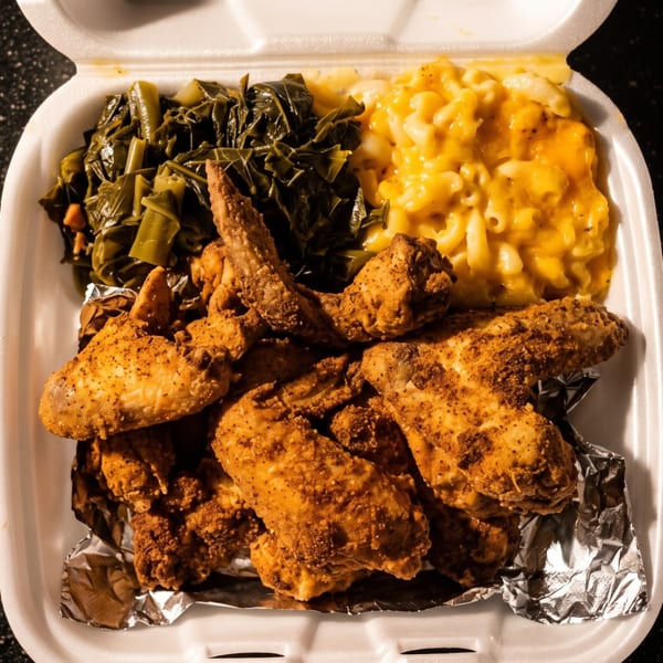 Fried chicken wings, collard greens, and macaroni and cheese in a white takeout container.