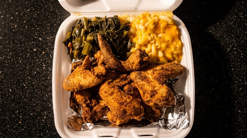 Fried chicken wings, collard greens, and macaroni and cheese in a white takeout container.